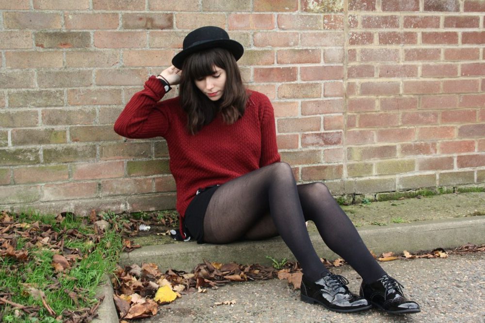 Black sheer tights with a burgundy knit sweater, black shorts, a bowler hat, and patent brogues