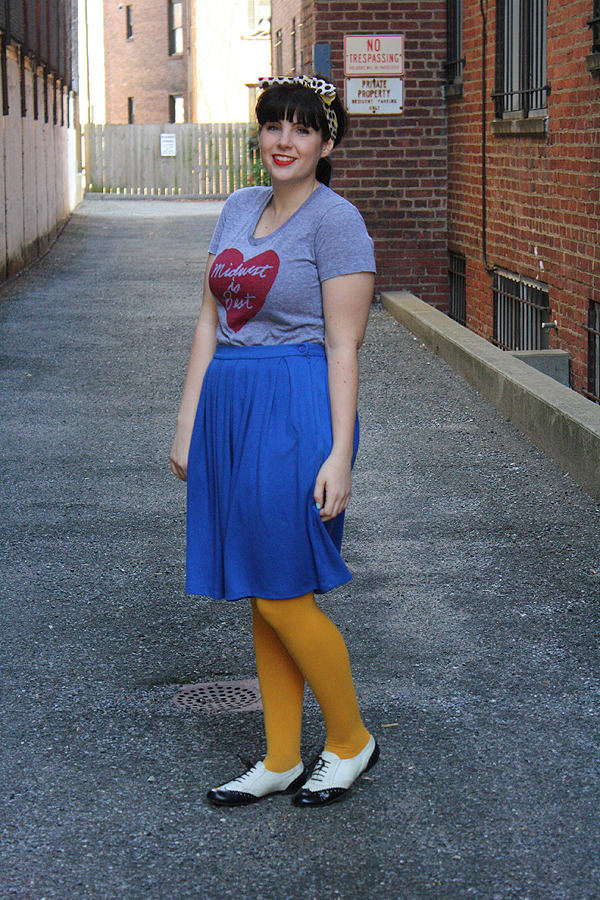 Mustard yellow tights with a bright blue pleated skirt, grey graphic tee, leopard print headband, and black and white oxford shoes