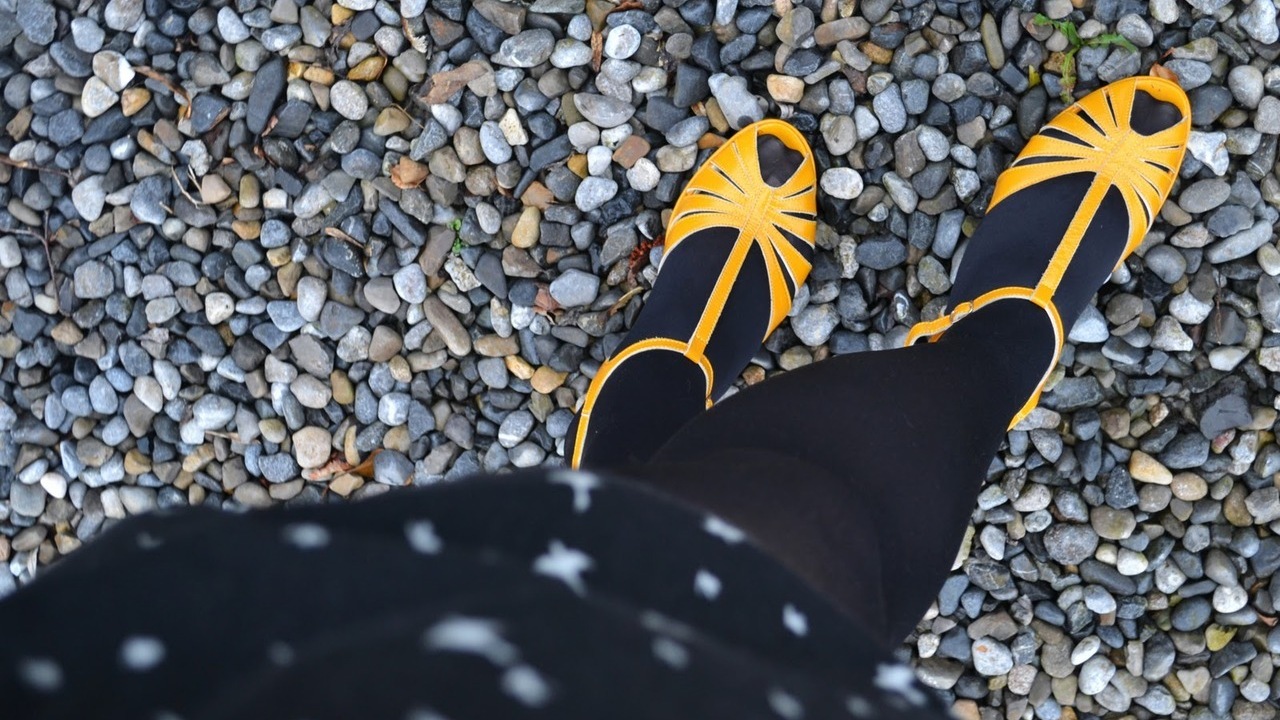 Feet in black tights and yellow strappy open toe sandals on pebbles, seen from above with a star-print skirt