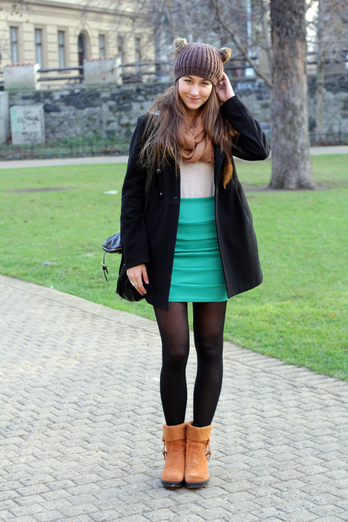 Black tights with light brown leather ankle boots, a teal skirt, a black coat and a knit beanie in a park