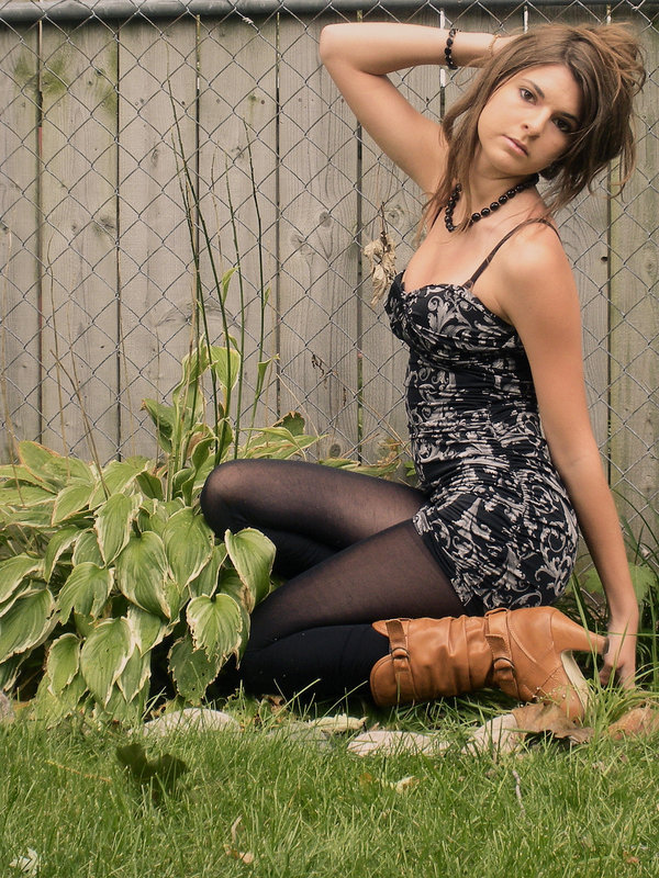 Black tights with slouchy cognac leather ankle boots and a fitted printed dress, sitting on grass by a fence