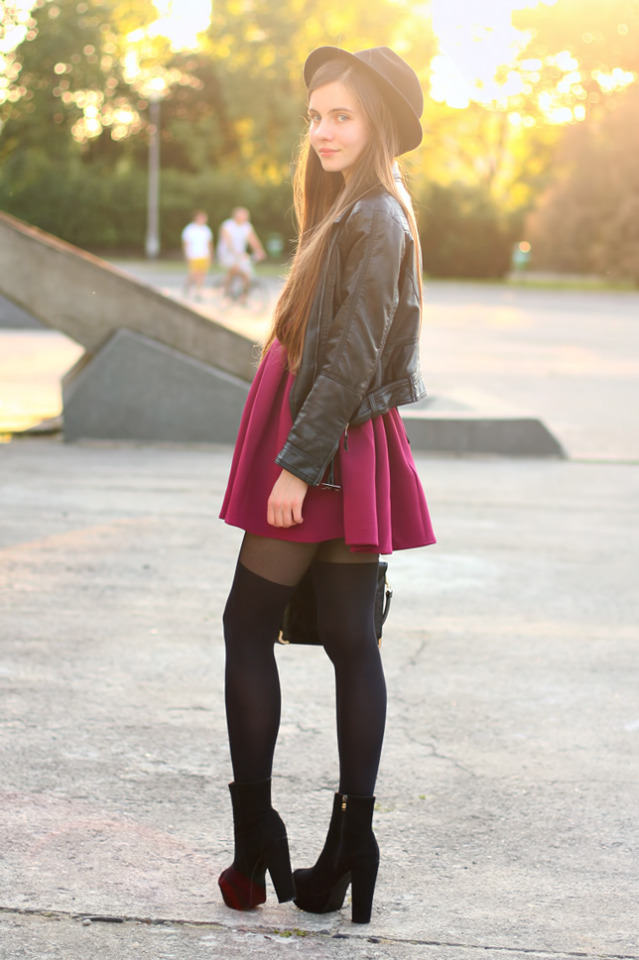 Woman in black faux thigh-high tights with a burgundy skater skirt and biker jacket, with a fedora and suede boots