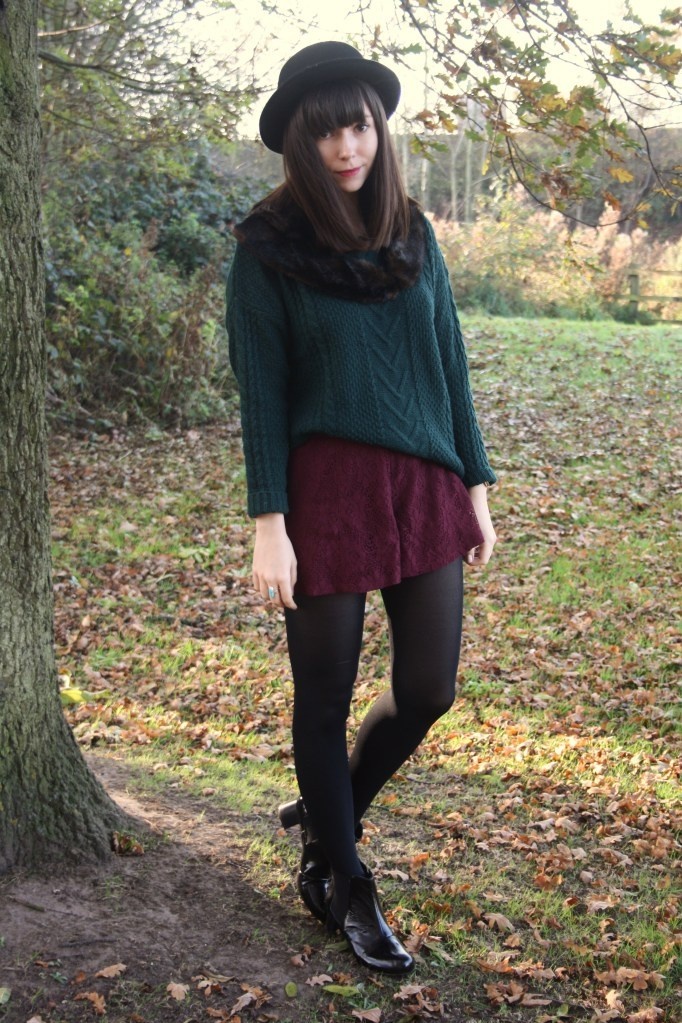Woman in black opaque tights with a burgundy skort and black ankle boots, wearing a dark green knit sweater and bowler hat in autumn leaves