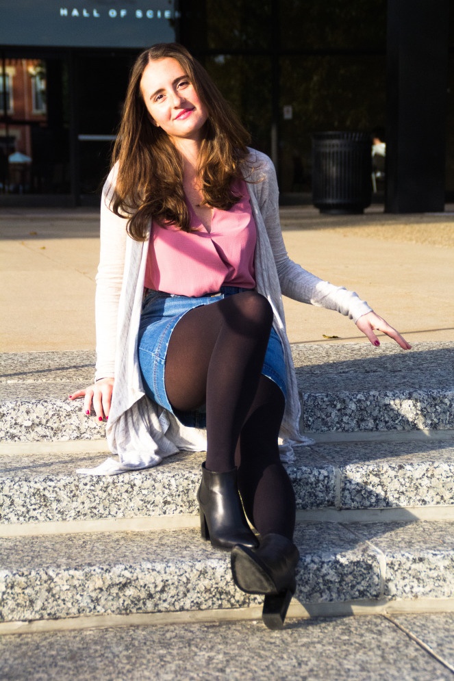 Woman in black opaque tights with a denim mini skirt and pink blouse, sitting on stone steps outside a building