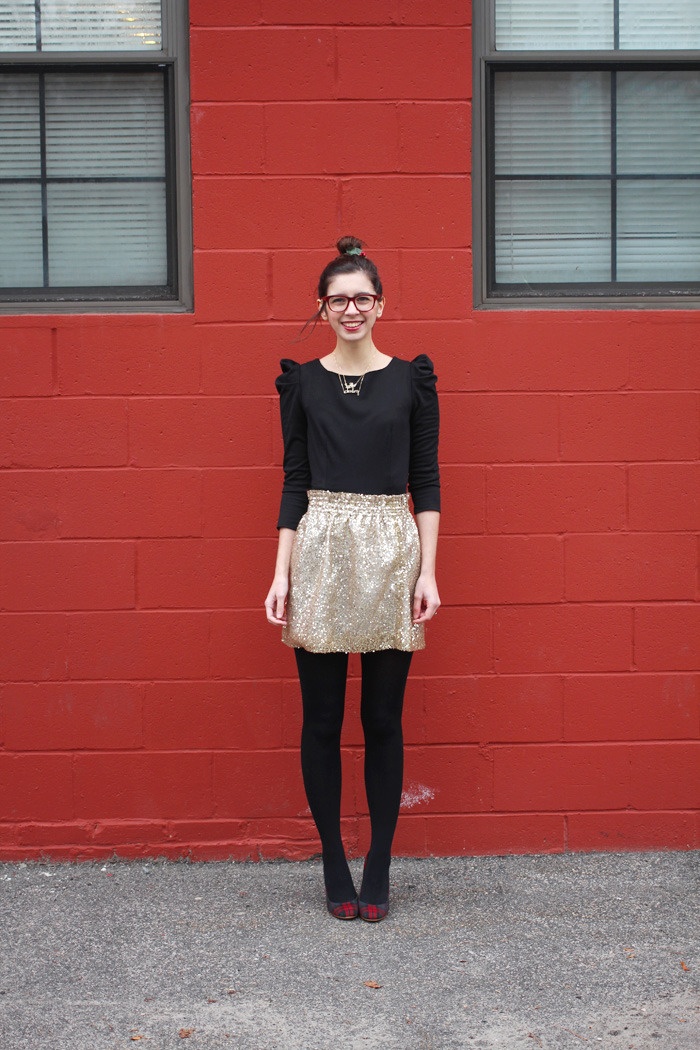 Woman in black opaque tights with a gold metallic mini skirt and black long-sleeve top, standing against a red brick wall