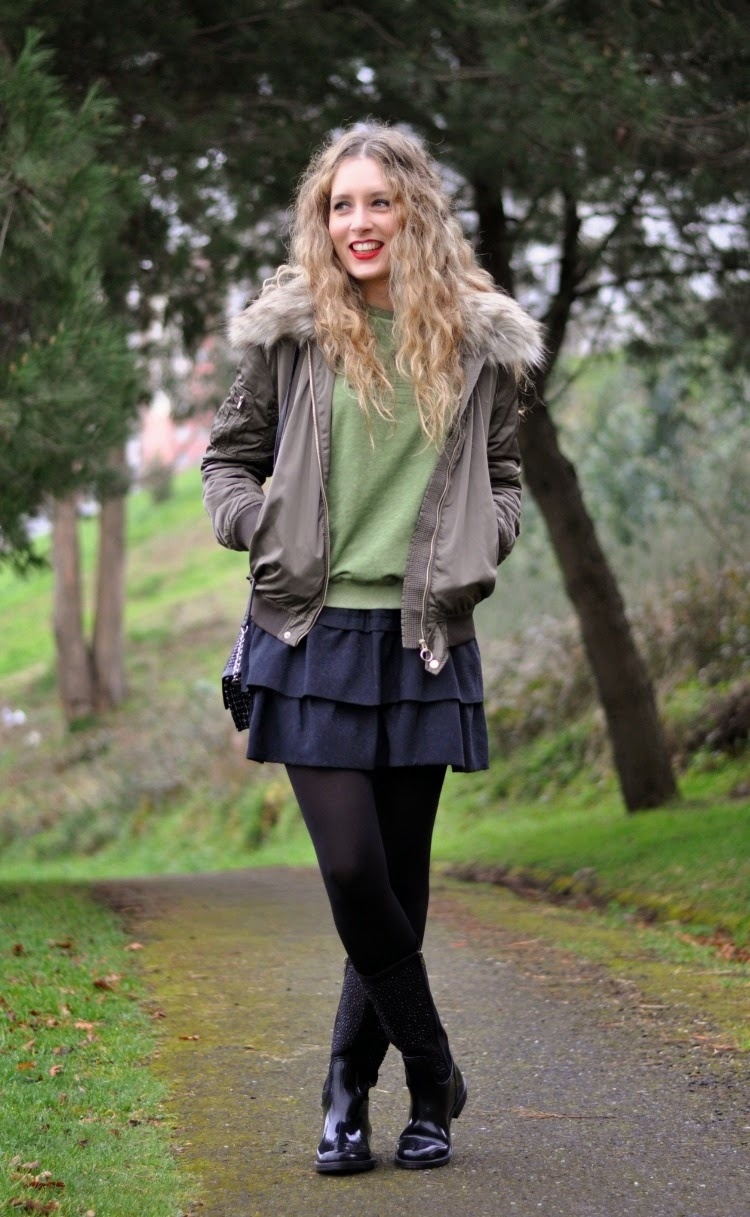 Woman in black opaque tights with a navy tiered mini skirt and olive green sweater, wearing a grey faux fur hood jacket on a park path