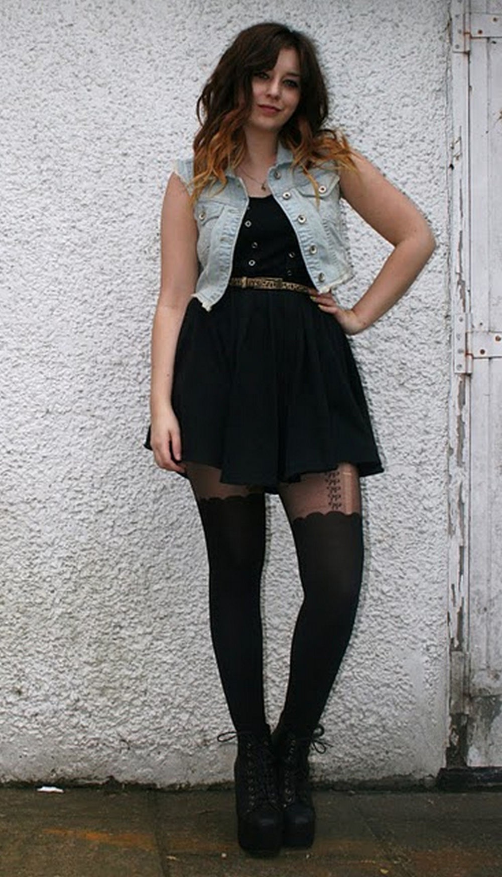 Woman in black tights with a faux stocking top and platform ankle boots, wearing a black skater dress and denim vest against a white wall