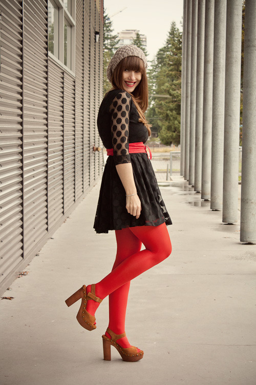 Woman in bright red tights with brown studded platform sandals and a black skirt, with a lace top and beanie