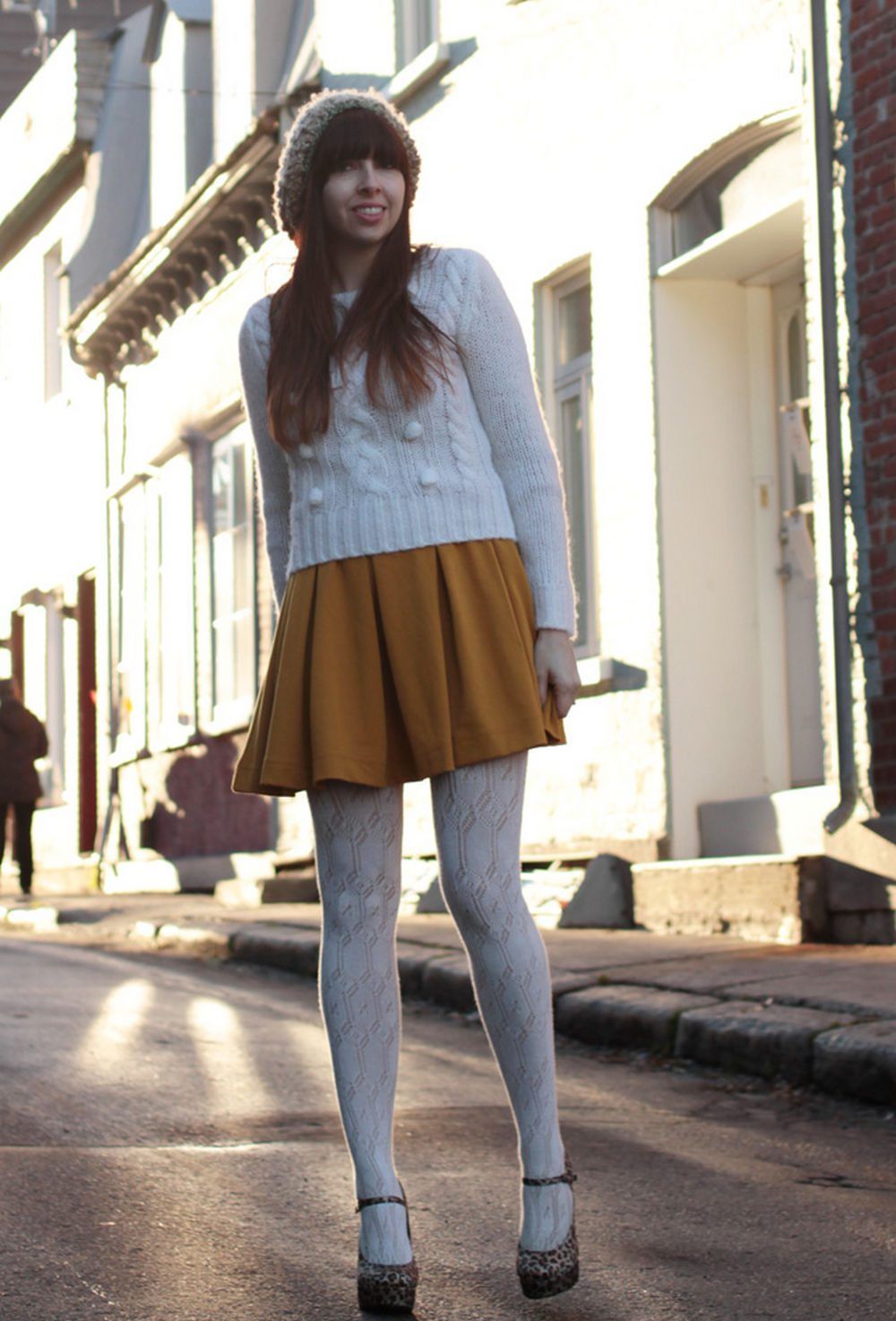 Woman in light grey lace tights with leopard print Mary Jane heels and a mustard pleated skirt, wearing a beanie