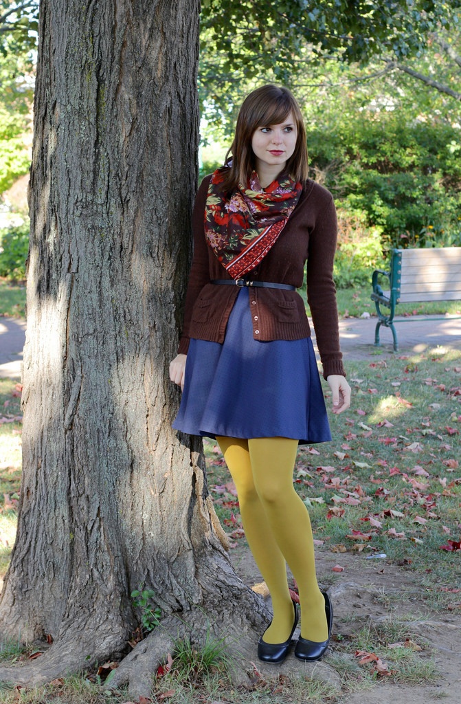 Woman in mustard yellow tights with a navy plaid mini skirt and brown cardigan, wearing a red scarf against a tree