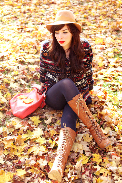 Woman in navy tights with cognac lace-up knee-high boots and a fair isle sweater, sitting in autumn leaves