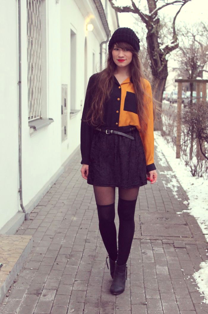 Woman in sheer black tights with black over-the-knee socks, wearing a lace skirt, an orange and black blouse and a beanie on a snowy street