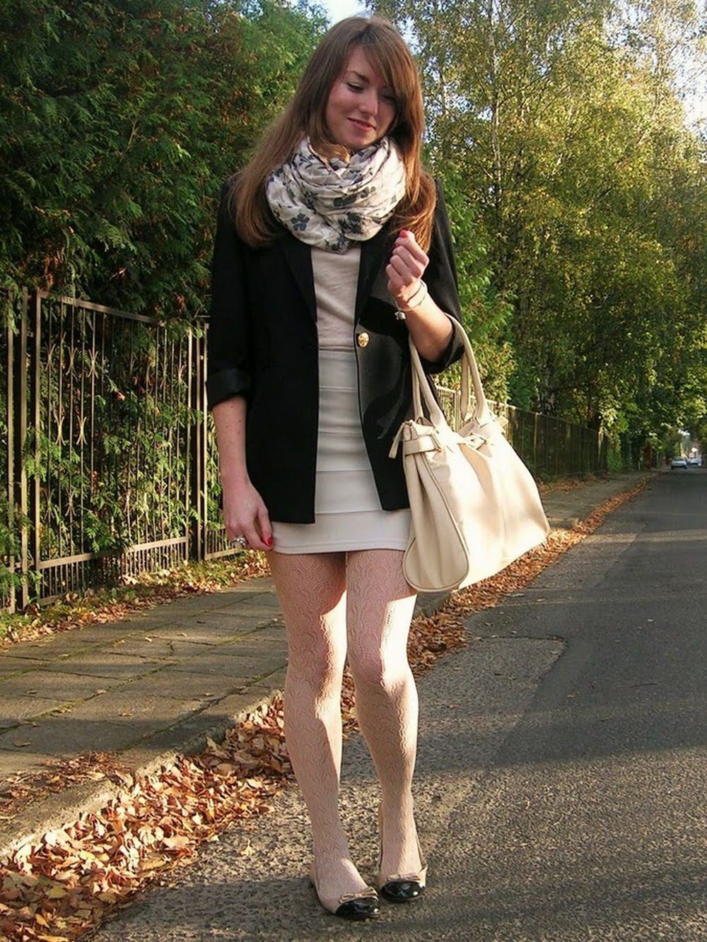 Woman in white fishnet tights with black and beige flats and a cream mini dress, wearing a black blazer on a leafy road