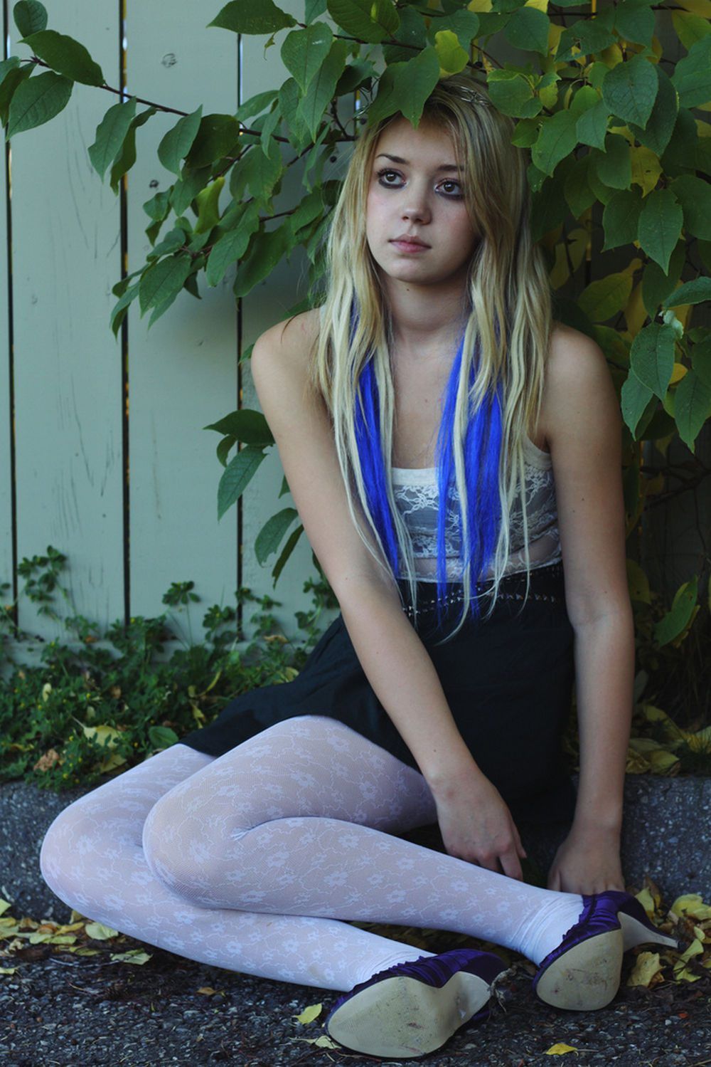 Woman in white heart pattern tights with purple ballet flats and a black skirt, sitting by a fence with blue hair extensions