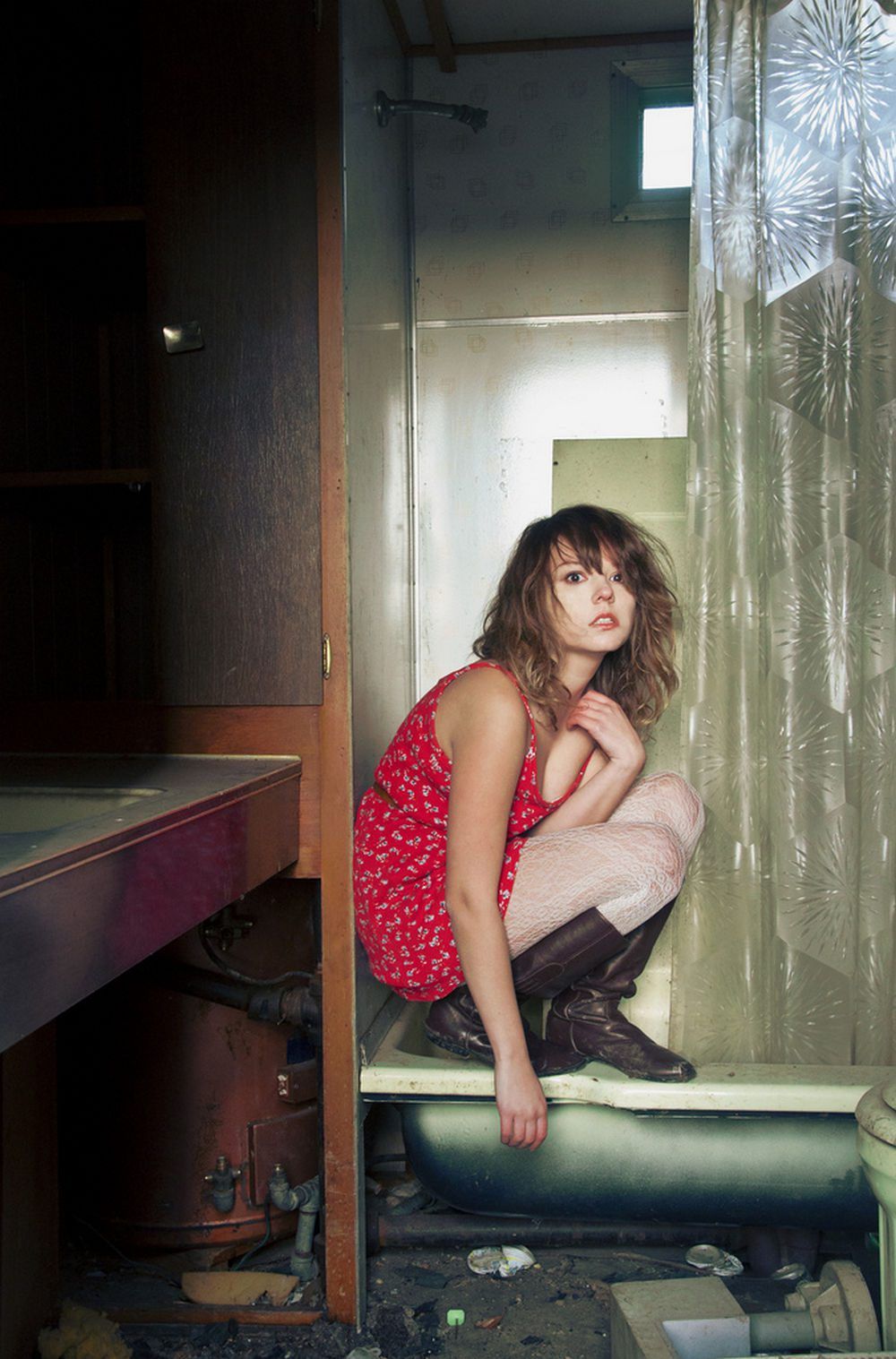 Woman in white lace tights with brown leather boots and a red floral dress, crouching in an old bathtub