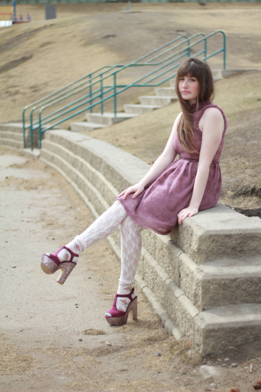 Woman in white lace tights with magenta glitter platform sandals and a dusty pink dress, sitting on a stone wall