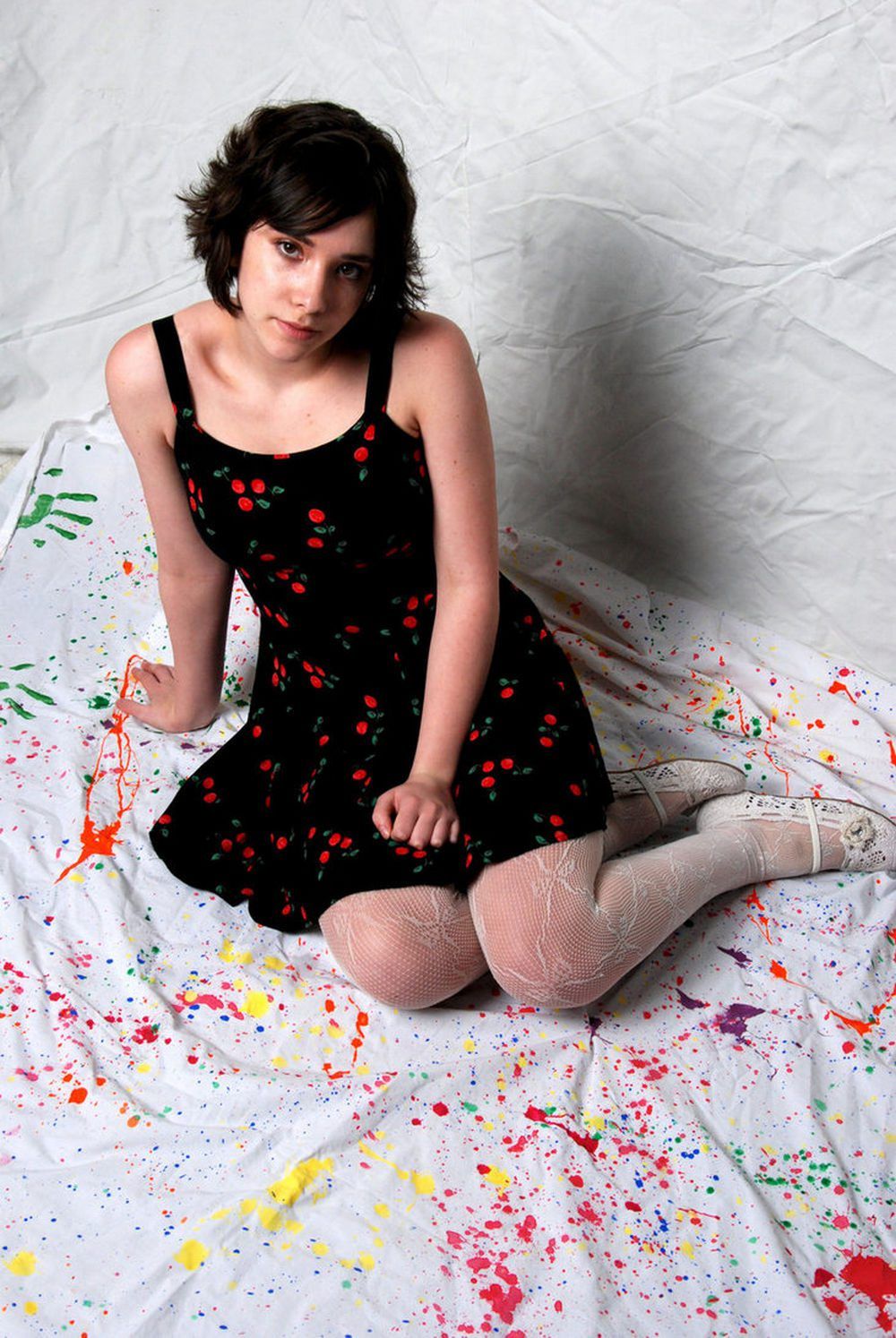Woman in white lace tights with white sneakers and a black cherry print dress, sitting on a paint splattered sheet