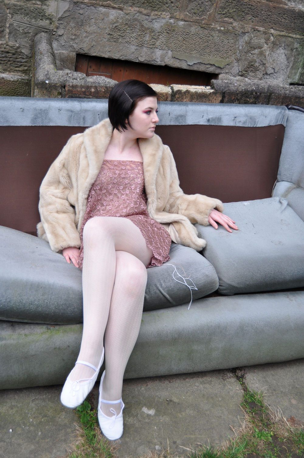 Woman in white sheer tights with white ballet flats and a dusty pink dress, wearing a faux fur jacket on an outdoor sofa
